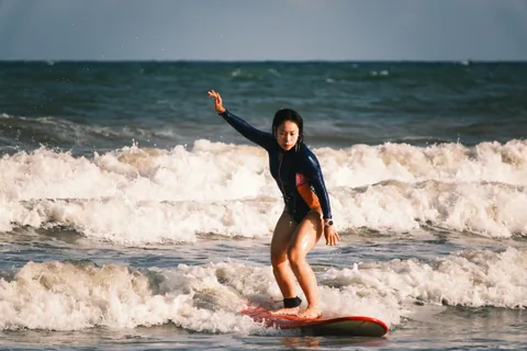 A student rides a red board through whitewater with one arm raised.