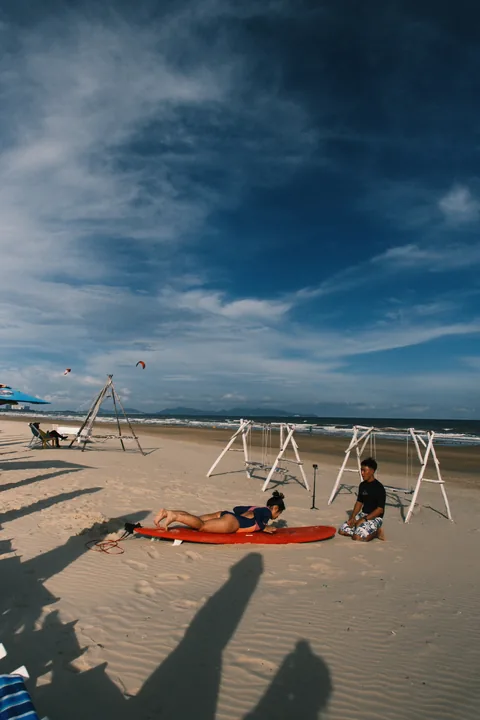 A student practises on a red surfboard on the sand with a coach nearby.
