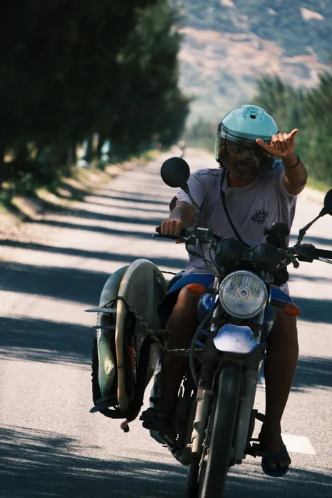 Surfers travel by motorbike with a board along a coastal road.
